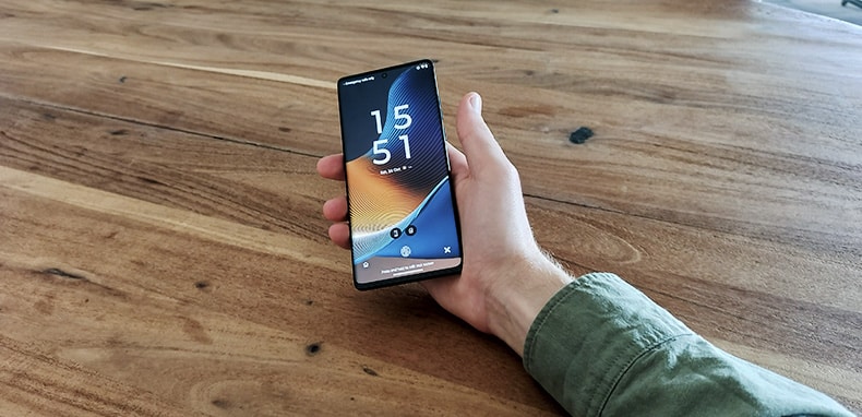 Een hand houdt de Motorola Edge 50 Fusion-smartphone vast, leunend op een houten tafel, het vergrendelscherm is zichtbaar. 